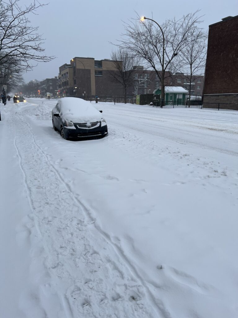 Car parked on the foreground. Sidewalk and street covered in thick snow. 